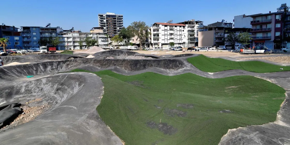 Bornova'da inşaatı devam eden, engebeli asfalt pisti ve yeşil suni çim alanlarıyla dikkat çeken bisiklet parkuru. Arka planda çok katlı apartmanların ve açık otopark alanının bulunduğu şehir manzarası yer alıyor.