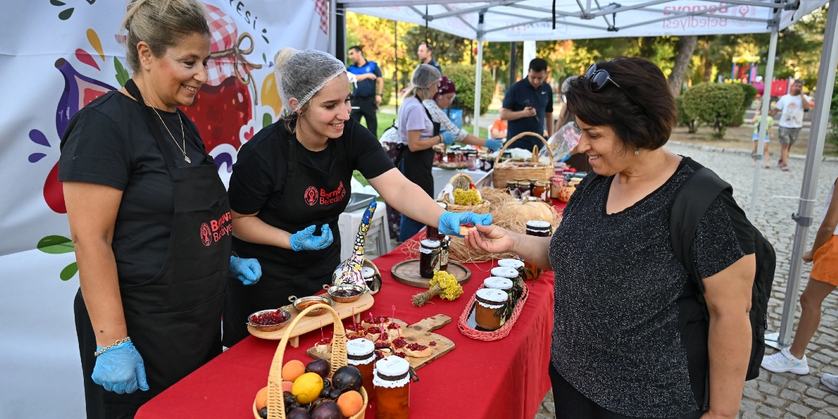 Bornova Belediyesi logolu siyah önlükler giymiş iki kadın, açık havada kurulan bir stantta ziyaretçilere doğal ürünlerden hazırlanan ikramları sunuyor. Mavi eldiven takan genç kadın, bir müşteriye üzerinde reçel bulunan küçük bir ekmek dilimi uzatıyor. Kırmızı örtülü masanın üzerinde çeşitli kavanozlarda reçeller, taze meyvelerle dolu bir sepet ve tadımlık ekmekler yer alıyor. Arka planda park ortamında yürüyen insanlar ve belediyeye ait beyaz bir çadır bulunuyor.