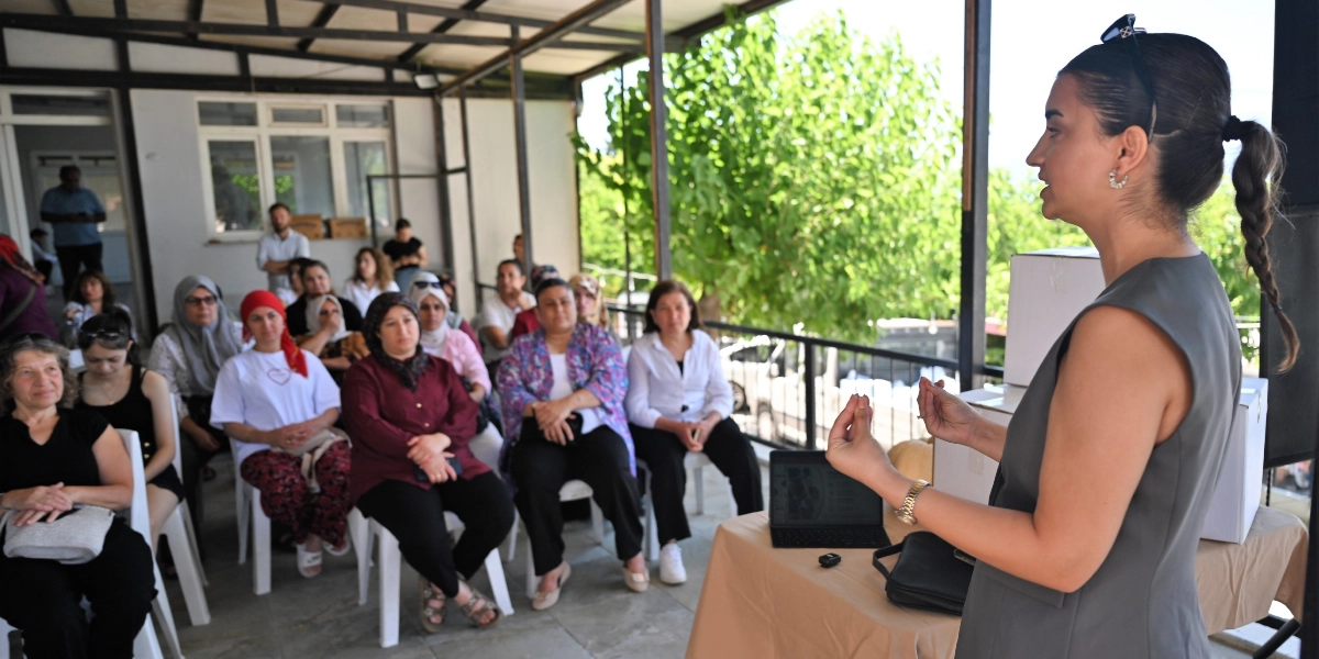 Açık havada, yarı kapalı bir alanda düzenlenen eğitim toplantısında, gri kolsuz bir bluz giyen genç bir kadın ayakta durarak katılımcılara sunum yapıyor. Kadının önündeki masada bir tablet bilgisayar bulunuyor. Karşısında oturan, çoğunluğu kadınlardan oluşan katılımcı grubu dikkatle konuşmacıyı dinliyor. Arka planda yeşil ağaçlar ve aydınlık bir ortam görülüyor.