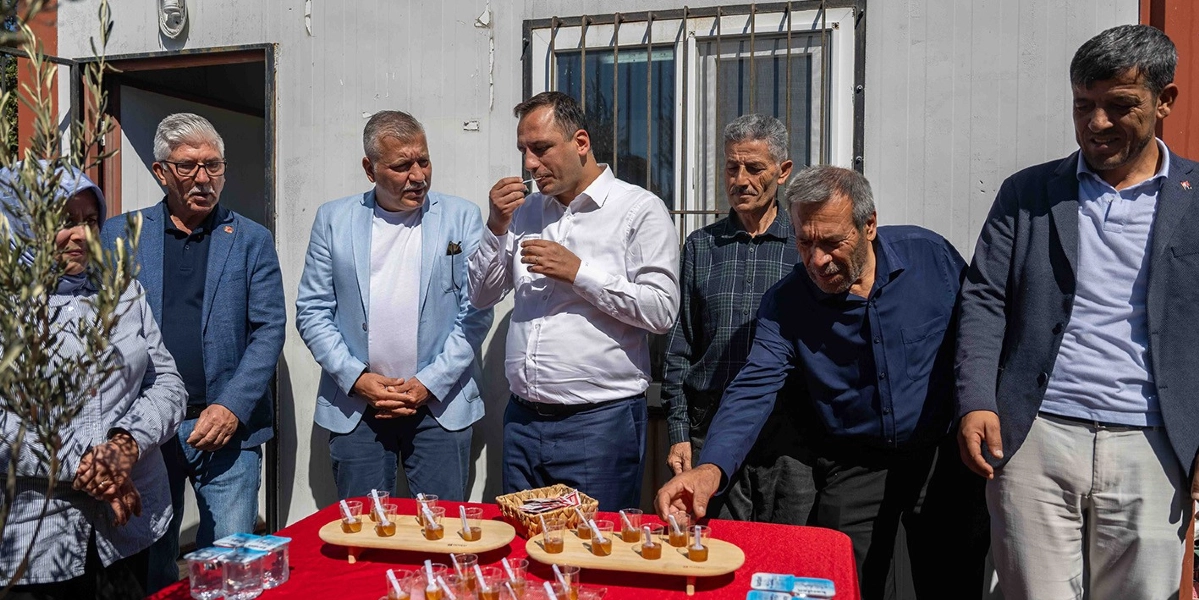 Açık havada, beyaz bir yapı önünde duran bir grup erkek ve bir kadın, kırmızı örtülü bir masanın etrafında toplanmış bal tadımı yapıyor. Masanın üzerinde ahşap sunumluklar içinde küçük plastik bardaklarda bal örnekleri bulunuyor. Gruptan beyaz gömlekli bir kişi elindeki bardaktan balı tadarken, yanındaki lacivert gömlekli bir kişi de masadaki bardaklardan birini işaret ediyor. Diğer kişiler ise meraklı ve gülümseyen bir ifadeyle onları izliyor.