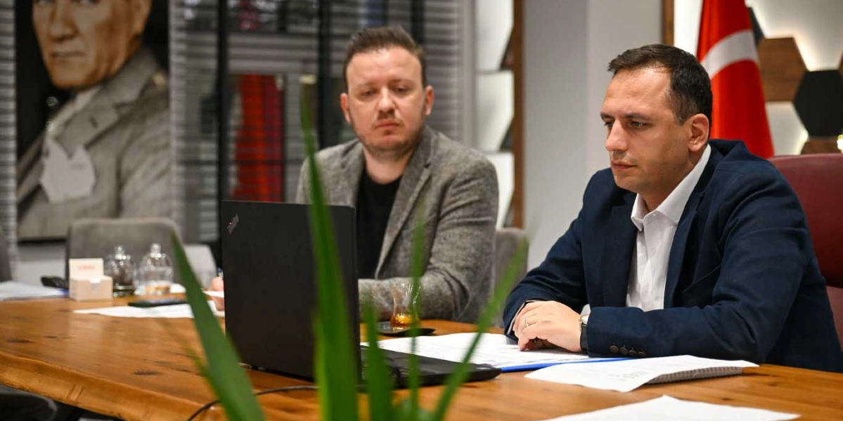 Ofis ortamında ahşap bir masada oturan iki erkek, önlerindeki dizüstü bilgisayara bakarak toplantı yapıyor. Sağ tarafta lacivert ceketli bir kişi, sol tarafta ise gri ceketli bir kişi yer alıyor. Arka planda duvarda Mustafa Kemal Atatürk'ün portresi ve Türk bayrağı bulunuyor.