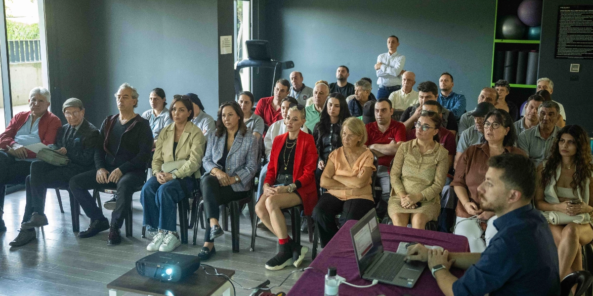 Modern bir spor salonu veya toplantı salonunda, bir sunumu dinleyen yaklaşık 30 kişilik bir grup. Ön planda, mor örtülü bir masada dizüstü bilgisayar başında oturan bir konuşmacı, arkasında ise sandalyelerde oturarak sunumu takip eden farklı yaş ve giyim tarzlarına sahip katılımcılar yer alıyor. Salonun arka duvarında spor ekipmanları ve bilgilendirme panoları bulunuyor.