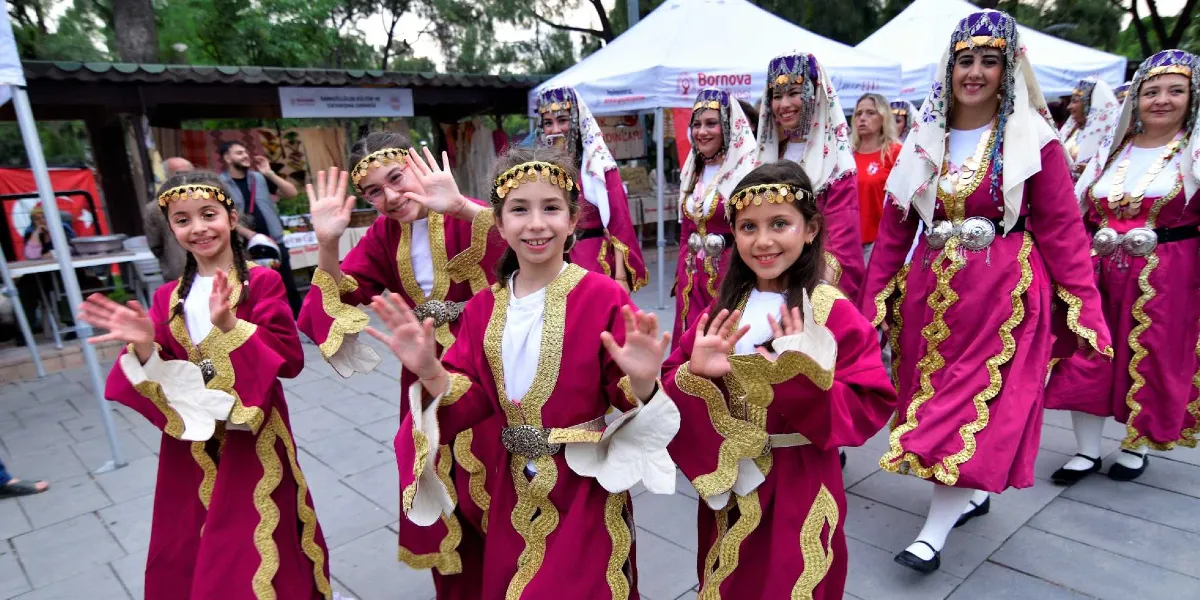 Geleneksel mor ve altın sarısı işlemeli kıyafetler giymiş, başlarında altın rengi süslemeler bulunan bir grup kız çocuğu ve kadın, açık havada neşeyle dans ederek ellerini havaya kaldırıyor. Arka planda Bornova Belediyesi logolu beyaz çadırlar ve etkinlik alanındaki stantlar görülüyor.