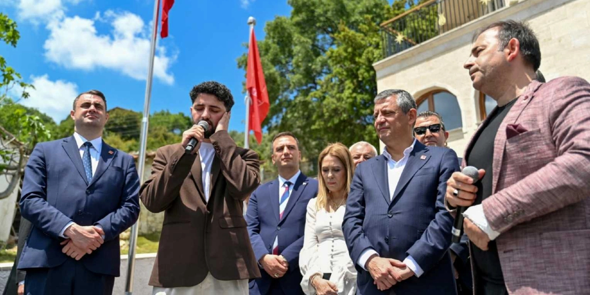CHP Genel Başkanı Özgür Özel ve beraberindeki heyet, açık havada düzenlenen bir törende yer alıyor. Mikrofonu tutan genç bir adam konuşma yaparken, Özgür Özel ve diğer katılımcılar dikkatle dinliyor. Arka planda Türk bayrakları, ağaçlar ve taş bir yapı görülüyor.