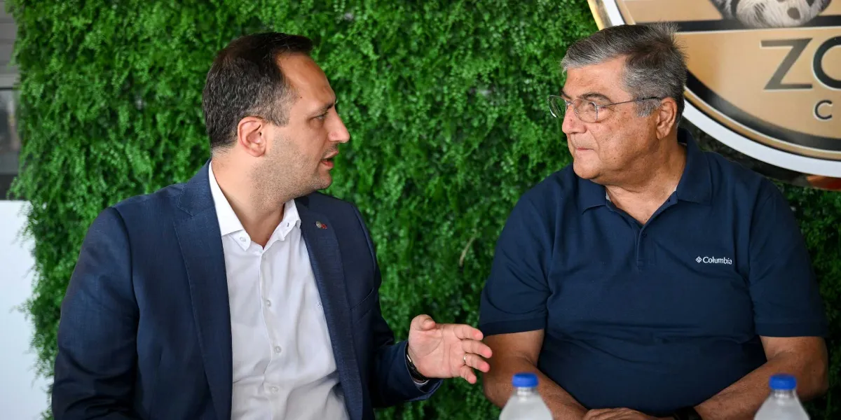 Koyu renk ceket ve beyaz gömlek giymiş bir erkek, yanında oturan gözlüklü ve lacivert polo yaka tişörtlü bir erkekle sohbet ediyor. Arka planda dikey yeşil yapraklı bir duvar dekoru ve kısmen görünen dairesel bir logo bulunuyor.