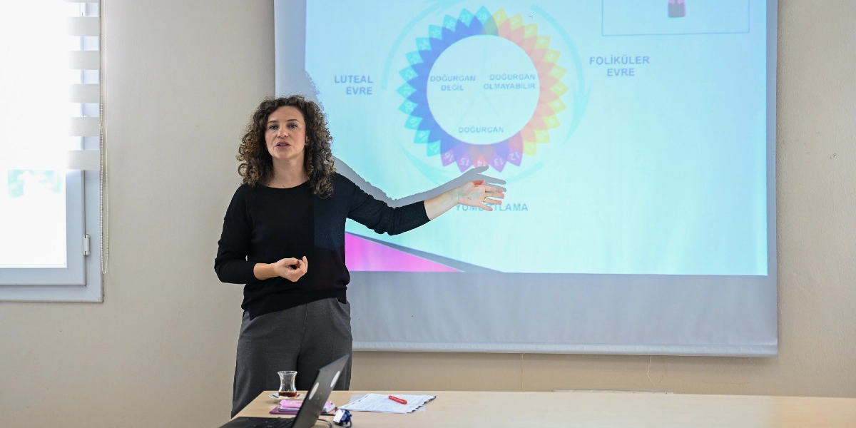 Kıvırcık saçlı bir kadın, bir sınıfta projeksiyon perdesinin önünde durarak sunum yapıyor. Kadın, elini perdede yer alan ve adet döngüsünü gösteren renkli bir şemaya doğru uzatmış durumda. Şemada 