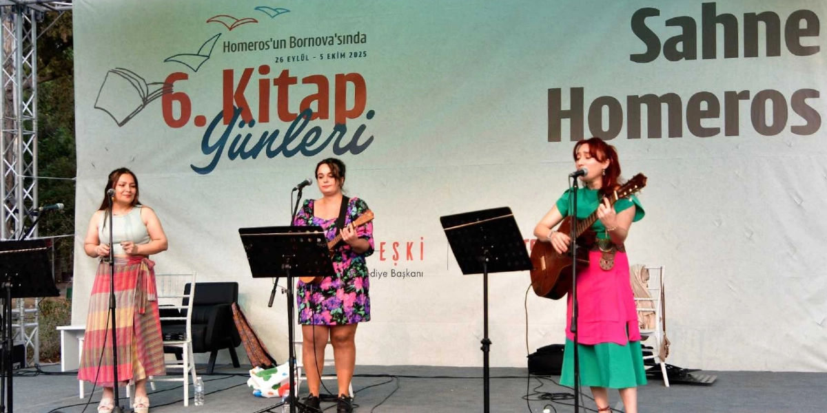 Homeros'un Bornova'sında 26 Eylül - 5 Ekim 2025 tarihleri arasında düzenlenen 6. Kitap Günleri etkinliğinin Sahne Homeros kısmında, üç kadın müzisyen sahnede performans sergiliyor. Soldaki kadın ayakta şarkı söylerken, ortadaki kadın ukulele, sağdaki kadın ise akustik gitar çalarak şarkıya eşlik ediyor. Arka planda etkinliğin adının ve tarihinin yazılı olduğu büyük bir branda yer alıyor.