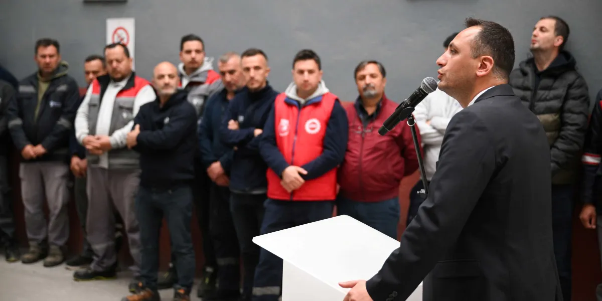 Ön planda takım elbiseli bir erkek, beyaz bir kürsü arkasında mikrofonla konuşma yapıyor. Arka planda ise iş kıyafetleri ve yelekler giymiş, dikkatle dinleyen bir grup erkek çalışan yer alıyor.