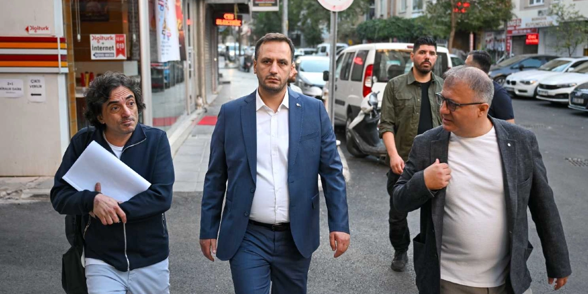 Bornova'da bir sokakta yan yana yürüyen üç erkek. Ortada lacivert takım elbiseli ve beyaz gömlekli bir kişi, solunda elinde beyaz bir dosya tutan koyu renk ceketli bir kişi, sağında ise gri ceket ve beyaz tişört giymiş gözlüklü bir kişi yer alıyor. Arka planda park halindeki araçlar ve iş yerleri görülüyor.