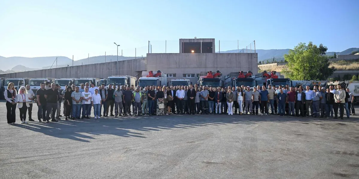 Geniş bir açık alanda, arkalarında sıralanmış çok sayıda iş makinesi ve kamyonun önünde yan yana duran, büyük çoğunluğu erkeklerden oluşan kalabalık bir grup çalışan. Arka planda beton bir duvar ve tepelik bir arazi görülüyor.