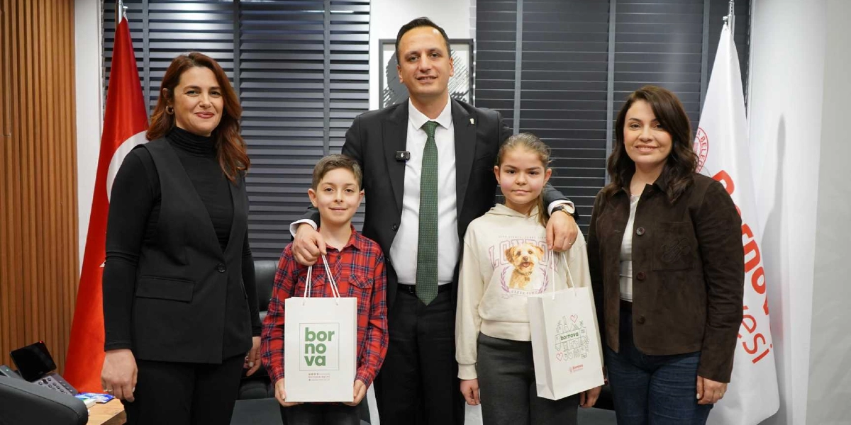 Bornova Belediye Başkanı, yanında iki kadınla birlikte ellerinde üzerinde Bornova Belediyesi logosu bulunan hediye poşetleri taşıyan iki öğrenciye eşlik ediyor. Arka planda Türk bayrağı ve Bornova Belediyesi flaması yer alıyor.