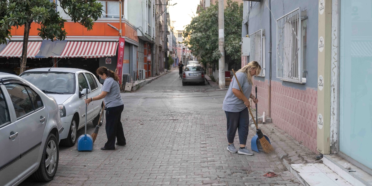 Gri tişörtlü ve koyu renk pantolonlu iki belediye temizlik görevlisi kadın, Bornova'da park halindeki araçların bulunduğu bir sokakta ellerinde süpürge ve faraşla sabah temizliği yapıyor.