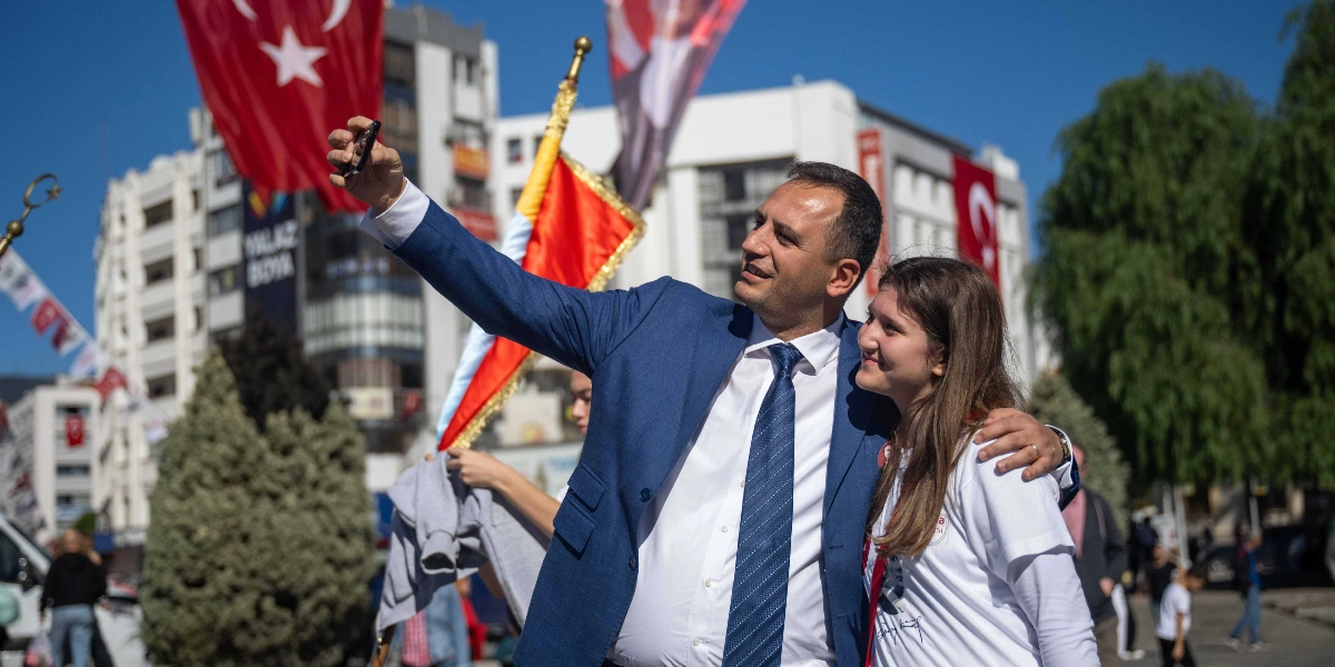 Mavi takım elbiseli ve kravatlı bir erkek, yanında duran genç bir kadınla birlikte dış mekanda selfie çekiyor. Arka planda Türk bayrakları, ağaçlar ve şehir merkezi binaları yer alıyor.