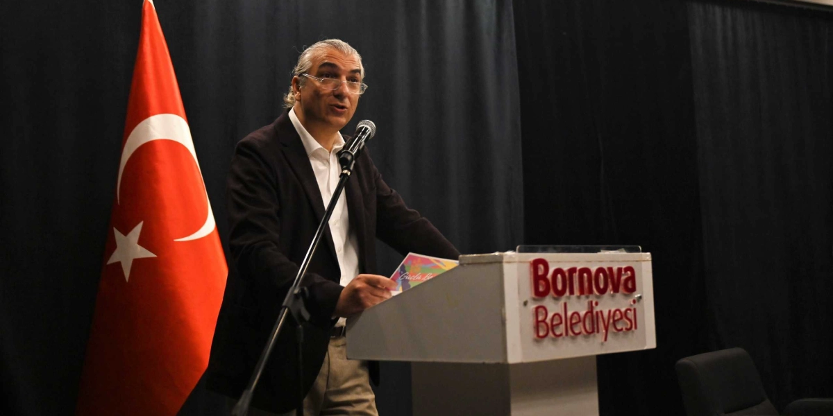 Koyu renkli bir perde önünde, üzerinde Bornova Belediyesi yazılı kürsüde konuşma yapan, gözlüklü ve kır saçlı bir erkek. Konuşmacının sol tarafında bir Türk bayrağı yer alıyor.