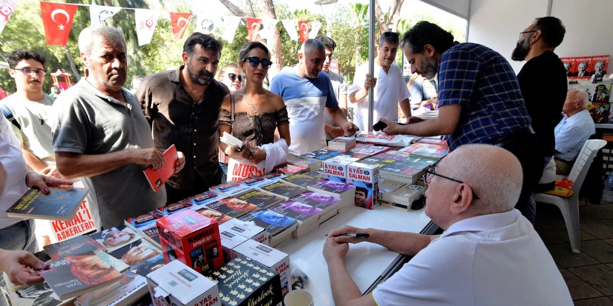 İlber Ortaylı, beyaz bir masanın arkasında oturmuş, önündeki kitapları imzalarken hayranlarıyla bir araya geliyor. Masanın üzerinde İlyas Salman’ın biyografi setleri ve çeşitli kitaplar dizili durumda. Ortaylı’nın karşısında, ellerinde kitaplar tutan ve imza sırası bekleyen farklı yaş gruplarından bir grup insan bulunuyor. Arka planda Türk bayrakları ve etkinlik alanını süsleyen flamalar yer alıyor.