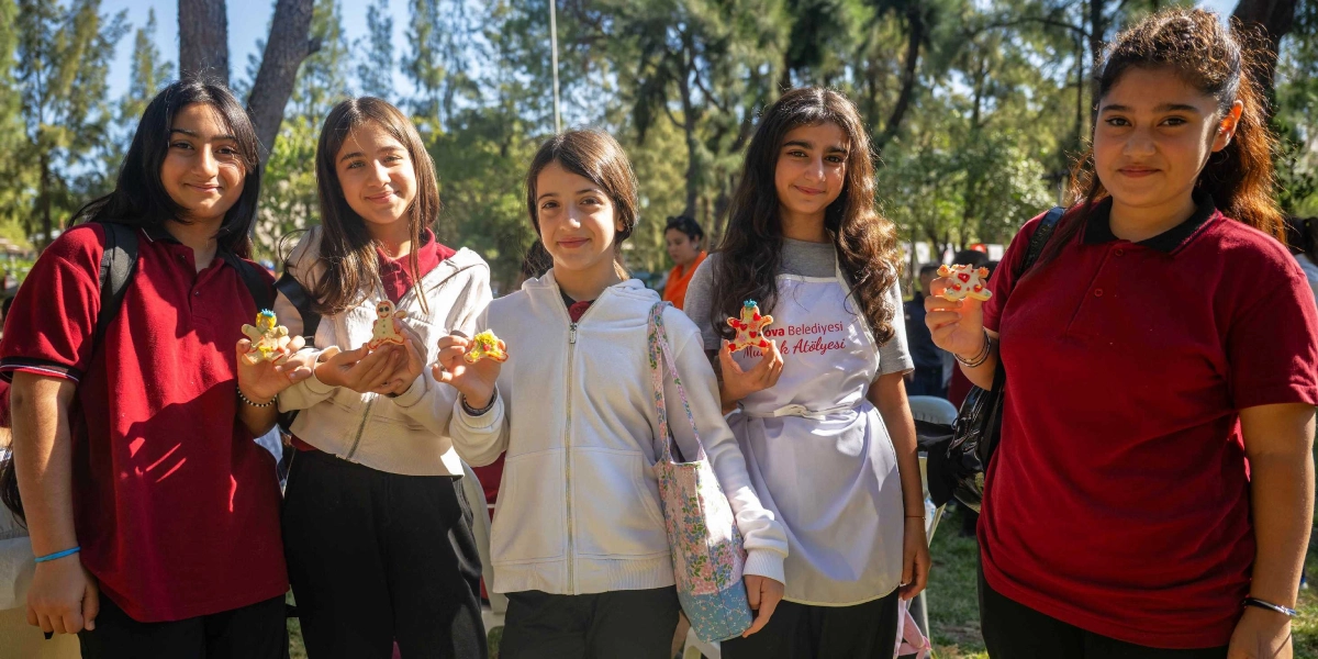 Açık havada, ağaçlık bir alanda yan yana duran beş genç kız, ellerinde süsledikleri kurabiyeleri göstererek gülümsüyor. Kızlardan biri üzerinde 