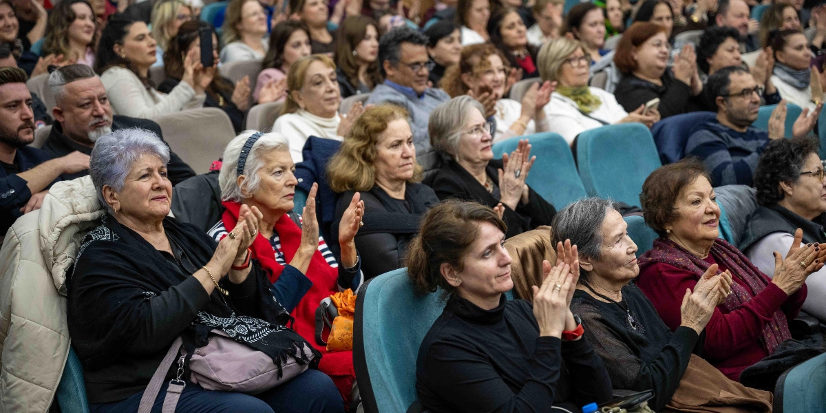 Konferans salonunda oturan ve sahnedeki bir performansı alkışlayarak izleyen, ağırlıklı olarak orta yaş ve üzeri kadınlardan oluşan kalabalık bir izleyici grubu.