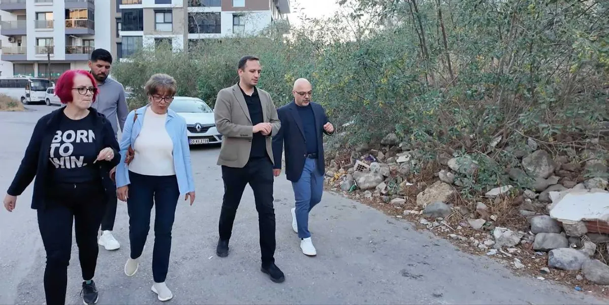 Bornova Belediye Başkanı Ömer Eşki ve beraberindeki heyet, bir sokak üzerinde yürüyerek incelemelerde bulunuyor. Başkan Eşki, üzerinde bej renkli bir ceket ve siyah bir tişörtle grubun önünde yer alıyor. Yanında açık mavi ceketli bir kadın, kırmızı saçlı bir kadın ve diğer yetkililer bulunuyor. Arka planda ise binalar, park halindeki araçlar ve taşlık bir arazi görülüyor.