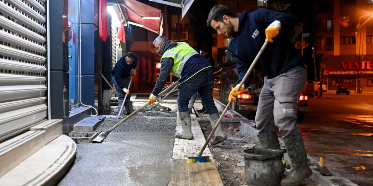 Gece vakti Bornova sokaklarında kaldırım yenileme çalışması yapan belediye işçileri, taze dökülen betonu mala ve süpürgelerle düzeltiyor. Üzerlerinde Bornova Belediyesi logolu kıyafetler, reflektörlü yelekler ve koruyucu çizmeler bulunan işçiler, dükkan önlerindeki kaldırım düzenlemesini tamamlamak için çalışıyor.