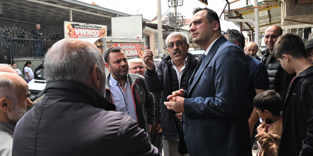 Koyu renk takım elbiseli bir erkek, sokak ortamında çevresindeki vatandaşlarla bir arada duruyor. Yanındaki gri saçlı ve gözlüklü bir adam el hareketleriyle konuşurken, takım elbiseli kişi onu dikkatle dinliyor. Çevrede başka erkekler ve bir çocuk da bulunuyor; arka planda ise üzerinde 