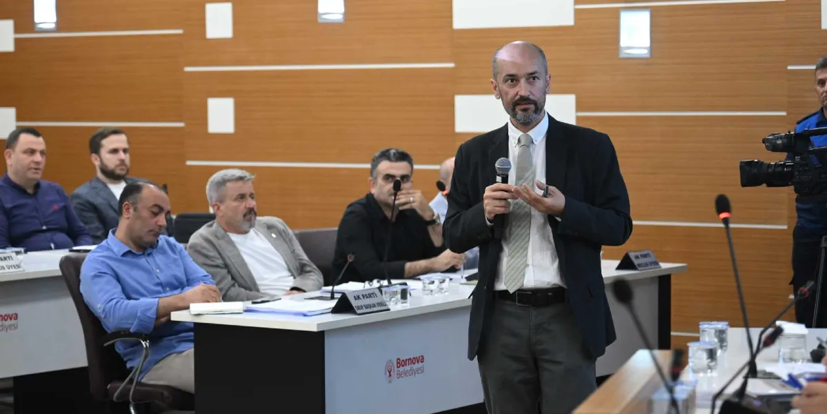 Bornova Belediyesi meclis salonunda, elinde mikrofonla konuşma yapan sakallı ve takım elbiseli bir erkek katılımcılara hitap ediyor. Konuşmacının arkasında ve yanındaki sıralarda oturan diğer meclis üyeleri onu dinliyor. Masaların üzerinde Bornova Belediyesi logoları ve AK Parti grup başkan vekili yazılı isimlikler yer alıyor. Sağ tarafta ise bir kameraman çekim yapıyor.