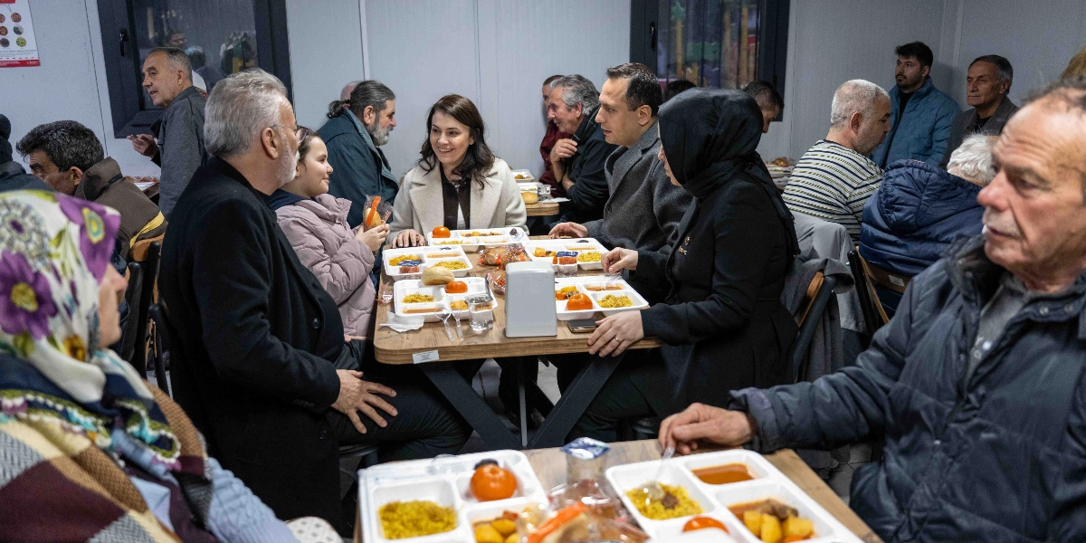 Konteyner kent veya benzeri bir toplu yaşam alanında, plastik bölmeli tabldot tabaklarda iftar yemeği yiyen çeşitli yaş gruplarından insanlar. Masalarda pilav, sulu yemek, domates ve ekmek bulunuyor. Ön planda başörtülü bir kadın ve yaşlı bir adam, orta planda ise gülümseyerek sohbet eden bir kadın ve çevresindeki diğer kişiler yer alıyor. Ortamda samimi bir atmosfer hakim.