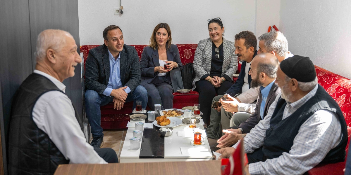 Kırmızı çiçek desenli bir koltukta yan yana oturan, aralarında yerel yöneticilerin de bulunduğu bir grup insan, önlerindeki çay ve atıştırmalıkların olduğu masanın etrafında sohbet ediyor. Grupta hem erkekler hem de kadınlar yer alırken, ortam samimi ve günlük bir ziyaret havası taşıyor.