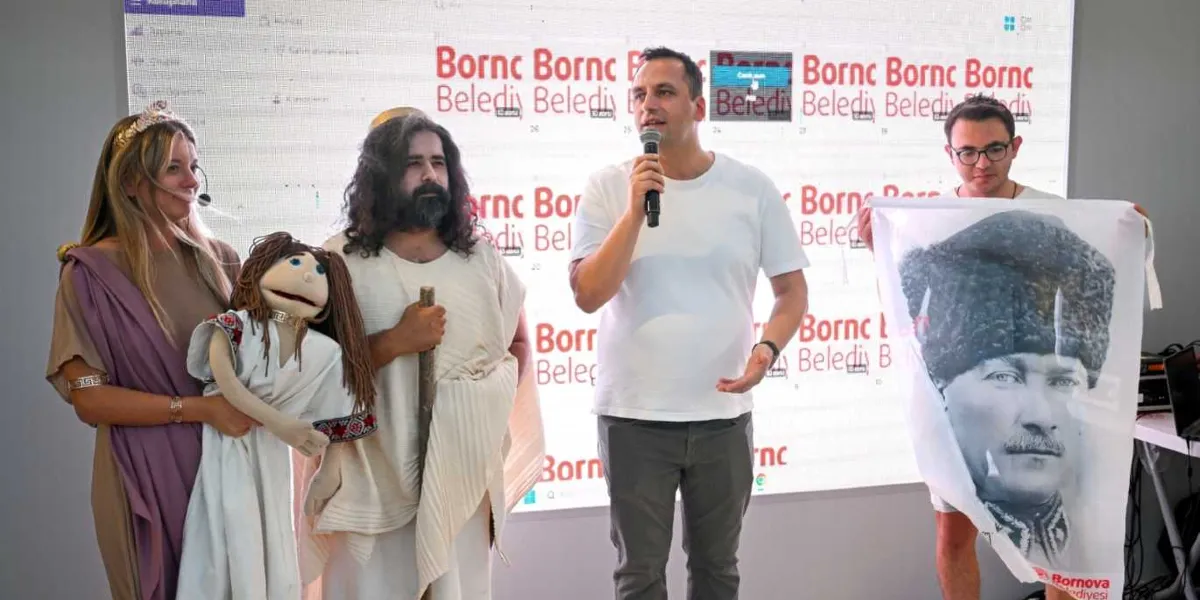 İzmir Enternasyonal Fuarı'ndaki Bornova Belediyesi standında, beyaz tişörtlü bir yetkili mikrofonla konuşma yaparken, yanında antik dönem kıyafetleri giymiş bir kadın ve elinde asasıyla uzun saçlı, sakallı bir erkek duruyor. Kadın elinde bir kukla tutarken, sağ tarafta gözlüklü bir genç, üzerinde Mustafa Kemal Atatürk'ün kalpaklı bir fotoğrafının bulunduğu beyaz bir bayrağı tutuyor. Arka planda üzerinde tekrarlayan 