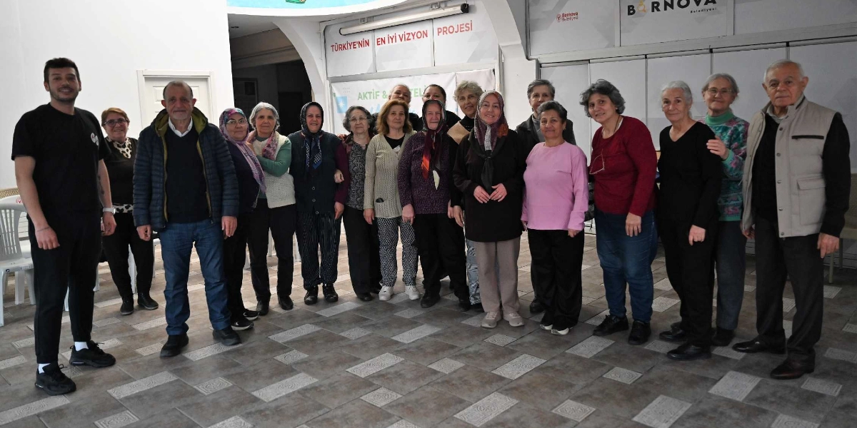 Bornova Belediyesi'nin düzenlediği bir etkinlikte, iç mekanda yan yana duran, çoğunluğu orta yaş ve üzeri kadınlardan oluşan yaklaşık 18 kişilik bir grup. Grubun en solunda genç bir erkek eğitmen, en sağında ise gri yelekli yaşlı bir beyefendi yer alıyor. Arka planda 