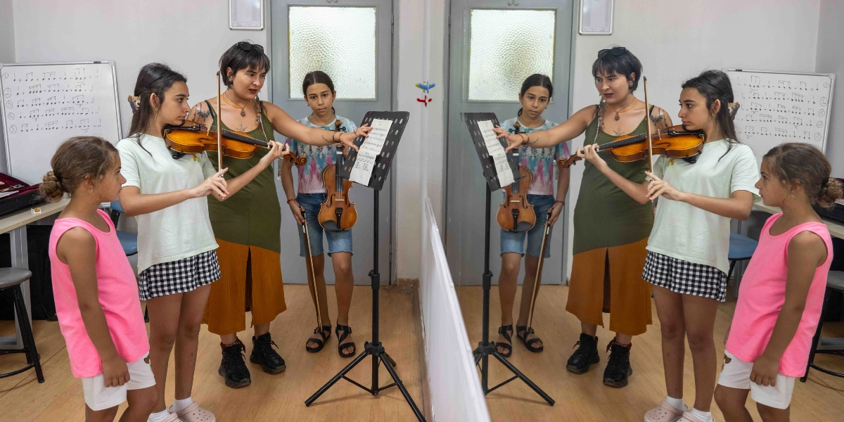 Müzik sınıfında bir eğitmen, elinde keman tutan genç bir öğrenciye nota sehpasındaki notaları göstererek ders veriyor. Eğitmen yeşil askılı bir elbise giymiş, öğrenci ise beyaz tişört ve ekose etekli. Yanlarında başka bir öğrenci daha bulunuyor, küçük bir kız çocuğu ise onları izliyor. Odanın ortasındaki büyük bir ayna, eğitmen ve öğrencilerin yansımasını oluşturarak sahneyi iki katına çıkarıyor. Arka planda üzerinde nota egzersizlerinin yazılı olduğu beyaz tahtalar yer alıyor.