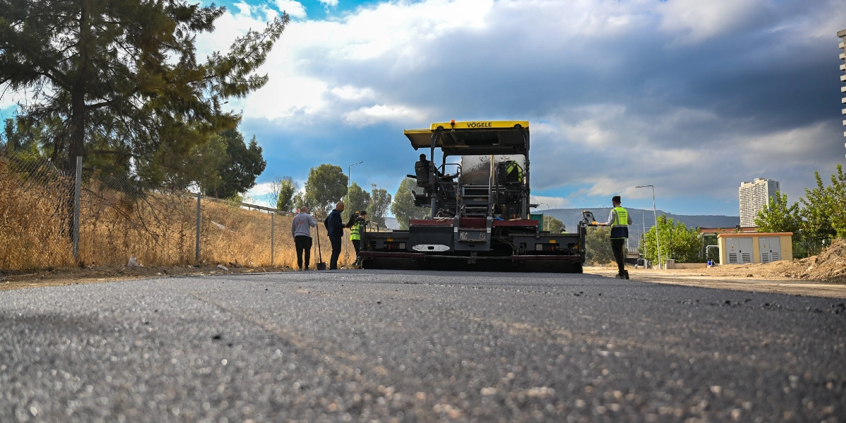 Açık havada, sarı renkli bir Vögele marka asfalt serim makinesinin yeni dökülmüş asfalt üzerinde çalıştığı yol yapım çalışması. İşçiler makinenin etrafında görevlerini yerine getirirken, arka planda ağaçlar, çitler ve gökyüzü görülüyor.