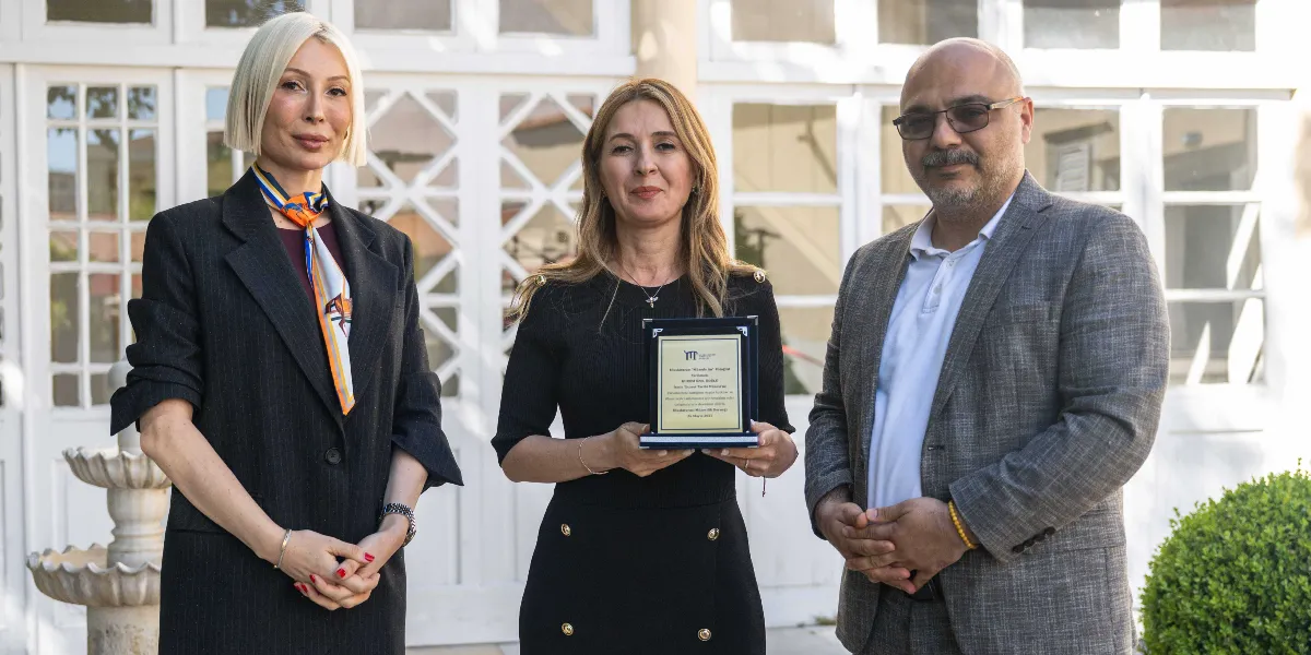 Beyaz, kafesli pencereleri olan tarihi bir binanın önünde duran üç kişi; solda sarı kısa saçlı, siyah ceketli bir kadın, ortada siyah elbiseli ve elinde plaket tutan bir kadın, sağda ise gözlüklü, gri ceketli bir erkek yer alıyor. Ortadaki kadının tuttuğu plaketin üzerinde 