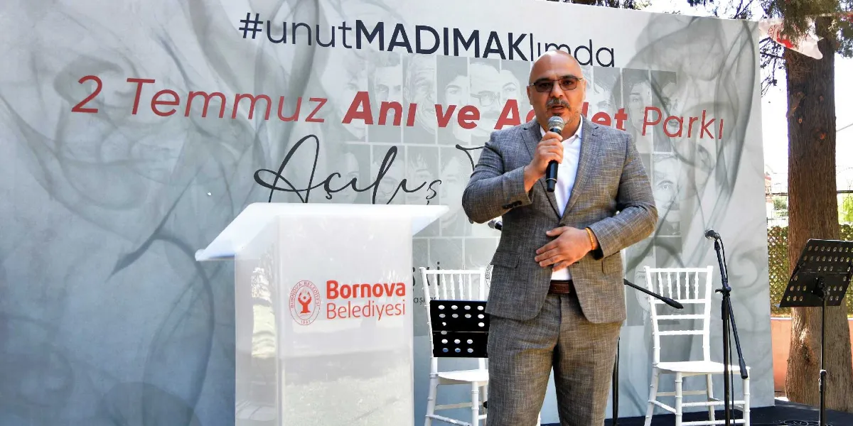Gri takım elbiseli, gözlüklü bir erkek, üzerinde Bornova Belediyesi logosu bulunan bir kürsünün önünde mikrofonla konuşma yapıyor. Arka planda, üzerinde duman efektleri, Madımak olayında hayatını kaybedenlerin silik fotoğrafları ile 