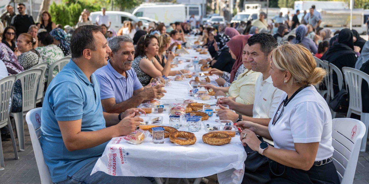 Açık havada, uzun bir masanın etrafında toplanmış kalabalık bir grup insan kahvaltı yapıyor. Masanın üzerinde simitler, küçük paketlerde kahvaltılıklar ve su bardakları bulunuyor. Ön planda, masanın bir ucunda oturan mavi tişörtlü bir erkek, yanında oturan gri saçlı bir adam ve karşılarında oturan beyaz tişörtlü bir kadın ile sohbet ediyor. Masanın üzerinde Bornova Belediyesi logosunun yer aldığı beyaz bir örtü serili. Arka planda ise masanın devamında oturan çok sayıda kişi yer alıyor.