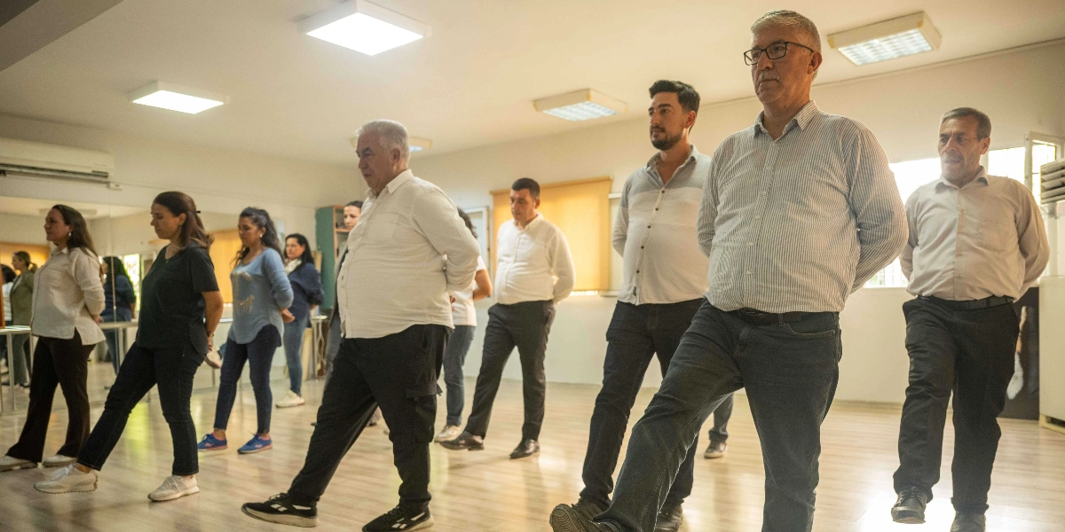 İç mekanda, geniş bir salonda yan yana dizilmiş bir grup kadın ve erkek, elleri arkalarında birleştirilmiş şekilde aynı yöne doğru adım atarak dans veya egzersiz çalışması yapıyor. Katılımcıların çoğu beyaz gömlek veya tişört giymiş durumda ve odaklanmış bir şekilde hareketleri takip ediyorlar.