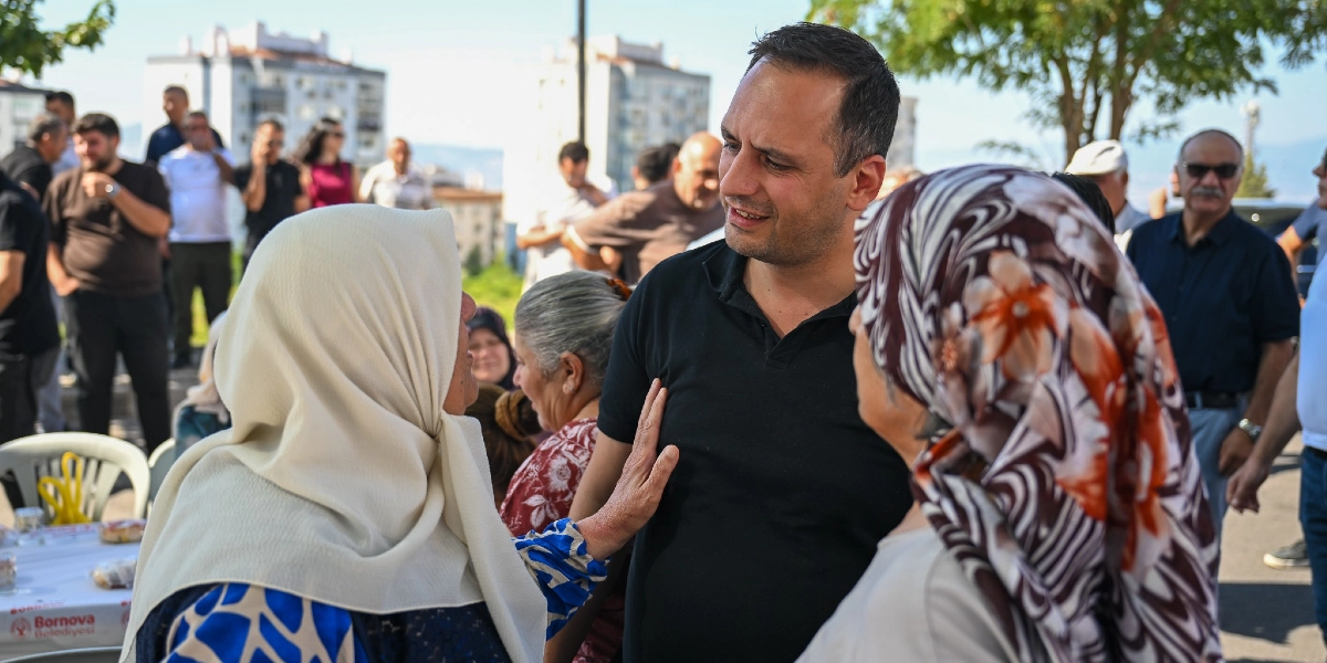 Siyah tişörtlü bir erkek, açık havada düzenlenen bir etkinlikte başörtülü kadınlarla sohbet ediyor. Kadınlardan biri elini samimi bir şekilde erkeğin omzuna koymuş, diğerleri ise onları dinliyor. Arka planda ise çeşitli insanlar ve binalar yer alıyor.