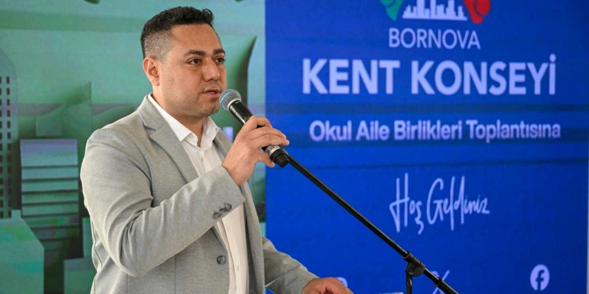 Gri ceketli ve beyaz gömlekli bir erkek, Bornova Kent Konseyi Okul Aile Birlikleri Toplantısı yazılı mavi bir fon önünde mikrofonla konuşma yapıyor.