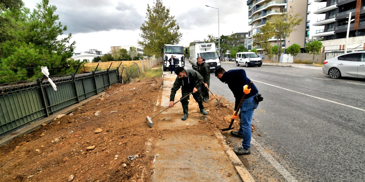 Belediye işçileri, yol kenarındaki toprak alanda kürek ve süpürge kullanarak çevre temizliği ve düzenleme çalışması yapıyor. İşçilerin arkasında park halindeki kamyonlar, sağ tarafta ise akan bir trafik yolu ve modern apartmanlar görülüyor.