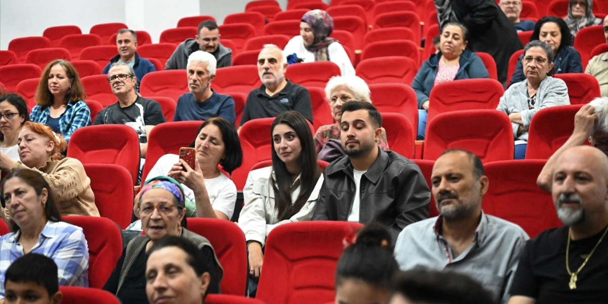 Kırmızı koltuklu bir konferans salonunda oturan ve sahnedeki etkinliği ilgiyle izleyen çeşitli yaş gruplarından izleyiciler. Ön sıralarda oturan bazı kişiler dikkatle sahneye bakarken, bir kadın elindeki telefonla çekim yapıyor.
