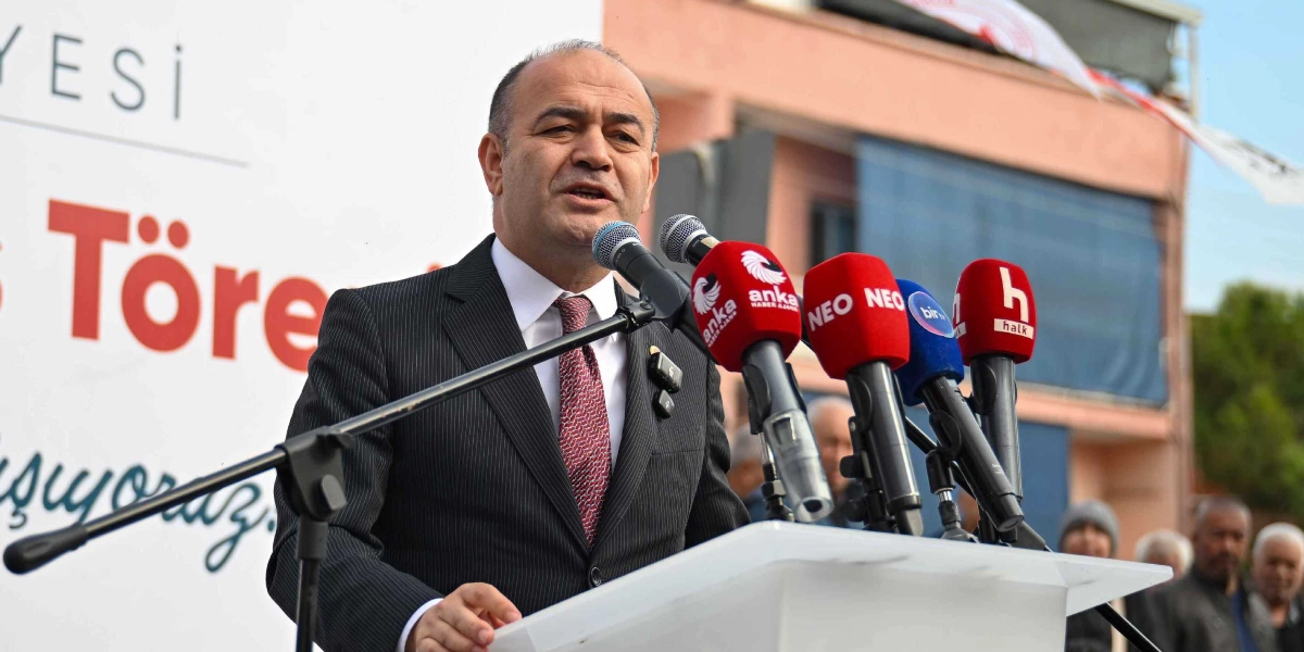 Koyu renk çizgili takım elbise ve kırmızı desenli kravat giymiş bir erkek, kürsüde konuşma yapıyor. Önünde Anka Haber Ajansı, NEO, Bir TV ve Halk TV mikrofonlarının bulunduğu bir mikrofon standı yer alıyor. Arka planda ise üzerinde 