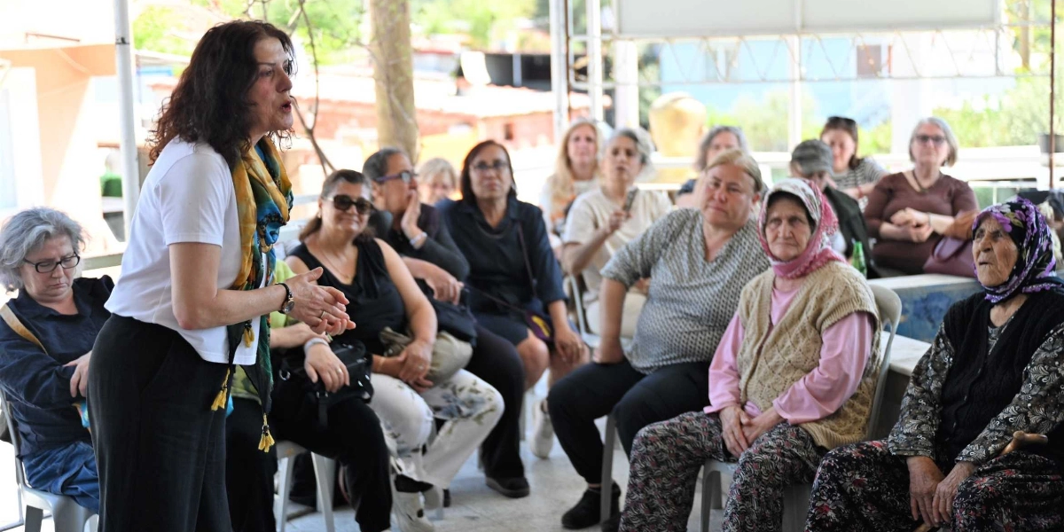 Açık havada düzenlenen bir toplantıda, beyaz tişörtlü ve renkli şallı bir kadın ayakta durarak konuşma yapıyor. Karşısında, farklı yaş gruplarından ve giyim tarzlarından oluşan, dikkatle dinleyen bir kadın grubu sandalyelerde oturuyor. Arka planda ise ağaçlar ve yerleşim yerleri görülüyor.