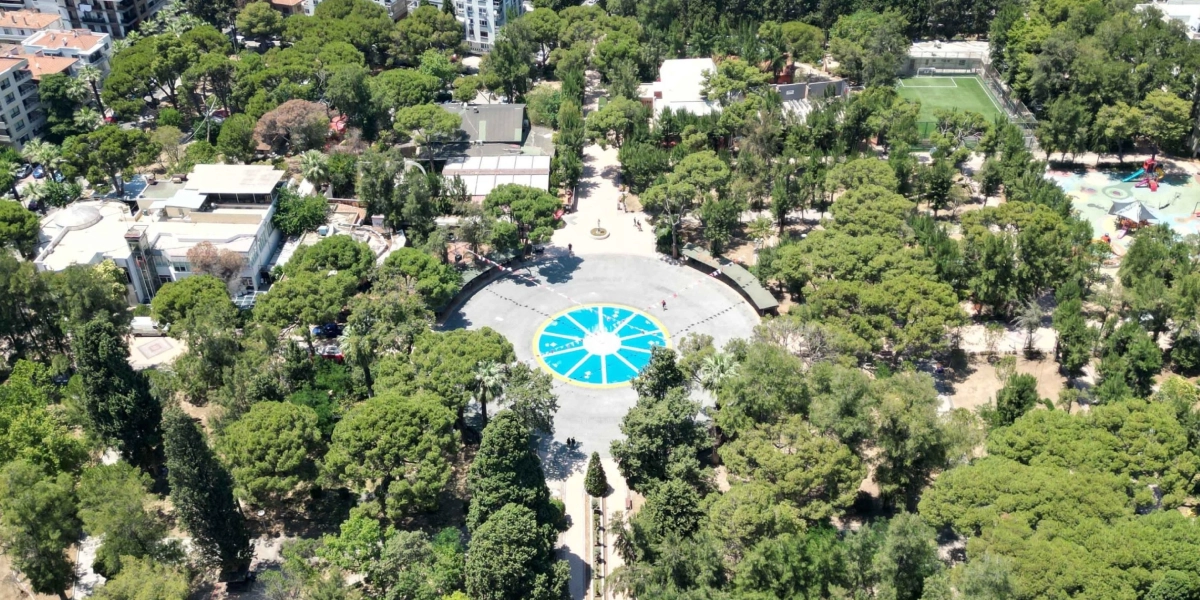 Bornova'da ağaçlarla çevrili geniş bir parkın kuş bakışı görünümü. Parkın merkezinde, üzerinde beyaz çizgilerle bölünmüş mavi renkli dairesel bir havuz veya zemin düzenlemesi yer alıyor. Parkın çevresinde yerleşim binaları, sağ üst köşede küçük bir futbol sahası ve çocuk oyun alanı bulunuyor.