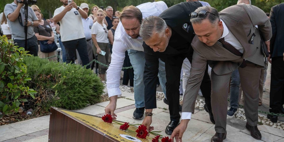 Üç erkek, açık havada bir anıtın üzerine kırmızı çiçekler bırakarak saygı duruşunda bulunuyor. Beyaz gömlekli, siyah ceketli ve gri takım elbiseli kişiler, eğilmiş bir şekilde anıta çiçekleri yerleştirirken, arka planda onları izleyen ve cep telefonlarıyla görüntü alan bir grup insan yer alıyor.