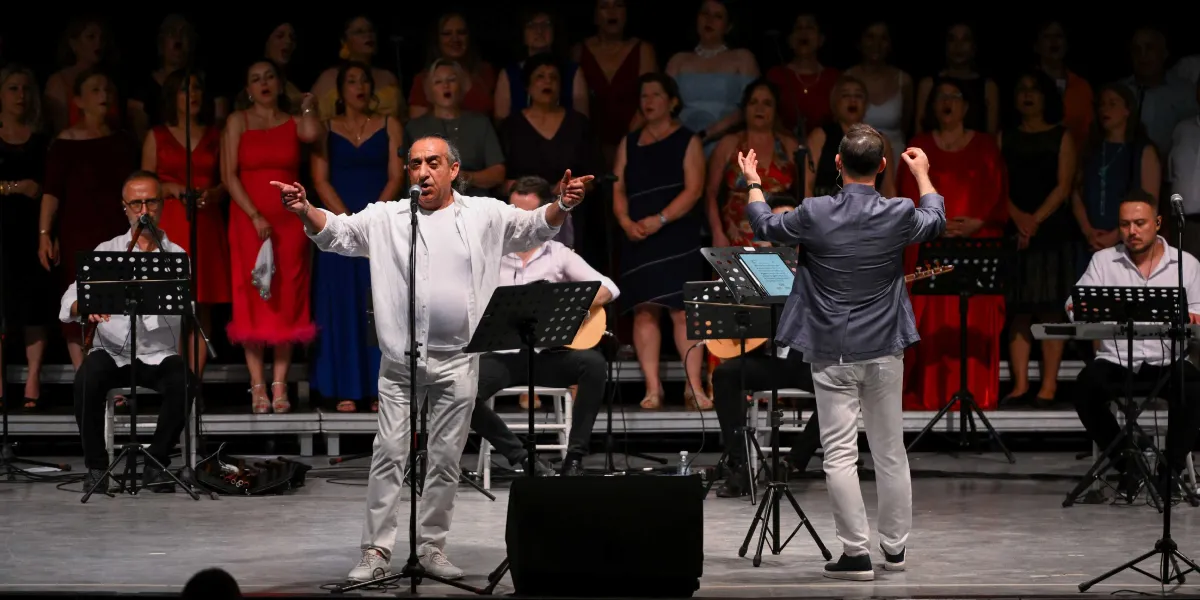 Sahne üzerinde beyaz kıyafetli bir solistin mikrofon başında şarkı söylediği, arkasında ise şefin yönetiminde çok sesli bir koronun yer aldığı türkü gecesi performansı. Solistin iki yanında enstrüman çalan müzisyenler bulunurken, koro üyeleri farklı renklerde kıyafetlerle sahne arkasında sıralanmış durumda.