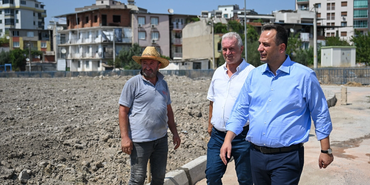 Hasır şapkalı bir çiftçi ile yanındaki iki kişi, Bornova'da tarım arazisi olarak hazırlanan toprak bir alanın kenarında yürüyerek incelemelerde bulunuyor; arka planda ise yerleşim yerleri ve apartmanlar görülüyor.