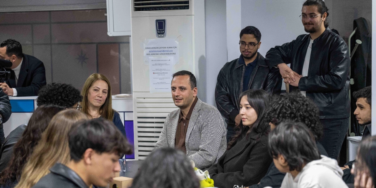 Kapalı bir mekanda bir masa etrafında toplanmış genç bir grup ve onlarla sohbet eden orta yaşlı bir erkek ile bir kadın. Arka planda beyaz bir klima ünitesi üzerinde, üzerinde e-posta adresi ve sosyal medya hesapları bulunan bilgilendirme yazıları asılı. Gruptaki gençler dikkatle dinlerken, ayakta duran iki genç erkek de konuşmaya dahil oluyor. Ortam samimi bir sohbet havasında geçiyor.