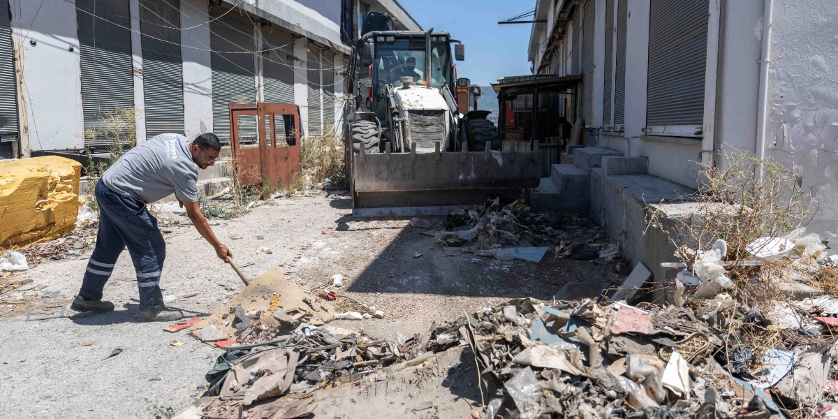 Bornova Belediyesi üniformalı bir görevli, iki bina arasındaki boşlukta biriken ayakkabı atıklarını ve çöpleri kürekle temizliyor. Görevlinin hemen arkasında, atıkları toplamak için bekleyen beyaz bir iş makinesi bulunuyor.