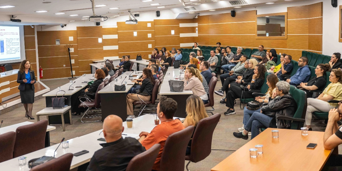 Modern bir konferans salonunda, kürsüde sunum yapan bir kadın konuşmacıyı dinleyen kalabalık bir katılımcı grubu. Salonun duvarları ahşap panellerle kaplı, katılımcılar ise kademeli olarak yerleştirilmiş yeşil koltuklarda oturuyor. Konuşmacının arkasındaki dev ekranda, üzerinde grafikler ve metinler bulunan bir sunum yansıtılıyor. Katılımcılar arasında farklı yaş ve giyim tarzlarına sahip kadın ve erkekler bulunuyor; bazıları not alıyor, bazıları ise dikkatle konuşmacıyı takip ediyor.