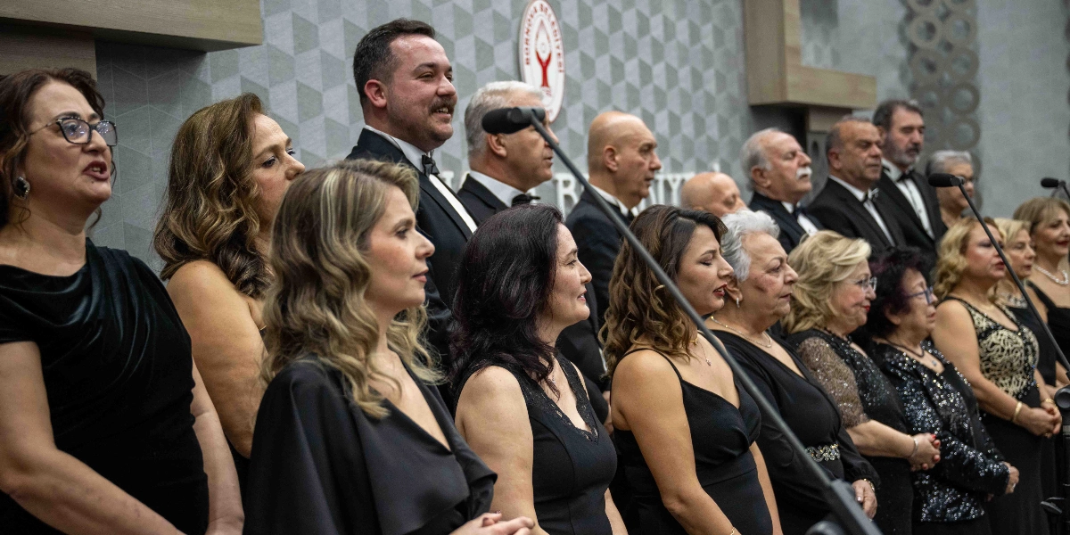 Resmi kıyafetler giymiş, çoğunluğu siyah elbiseli kadınlar ve takım elbiseli erkeklerden oluşan bir koro, Bornova Belediyesi logosunun bulunduğu bir arka plan önünde şarkı söylüyor. Koro üyeleri yan yana dizilmiş, odaklanmış bir şekilde ileriye bakıyor ve önlerinde mikrofonlar bulunuyor.