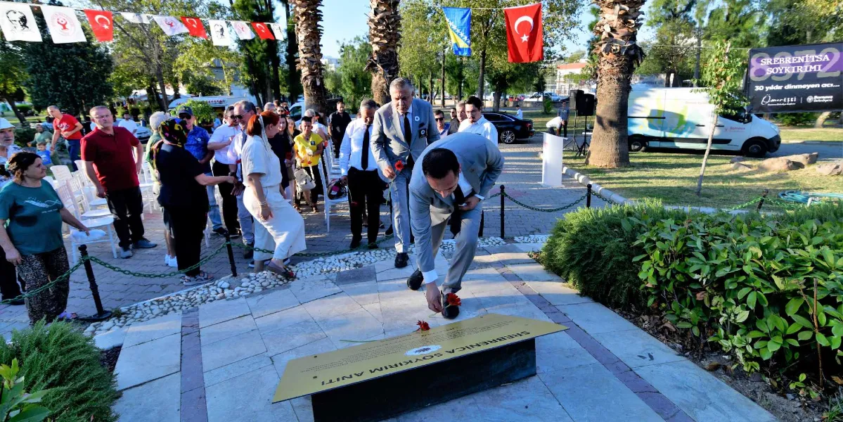 Bornova'da düzenlenen Srebrenica Soykırımı anma töreninde, takım elbiseli bir yetkili park içerisindeki anıt plaketine kırmızı bir karanfil bırakıyor. Arka planda törene katılan bir grup insan, Türk bayrakları ve Bosna-Hersek bayrakları altında sıralanmış sandalyelerde oturuyor veya ayakta bekliyor. Sağ tarafta üzerinde 